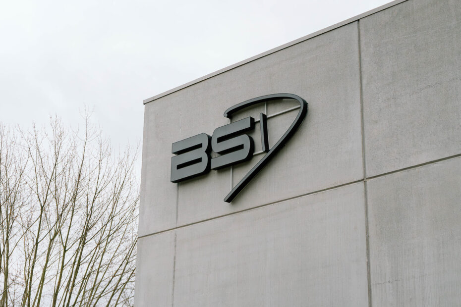 Zwart BSI-logo bevestigd aan een grijze muur van een gebouw met bomen op de achtergrond.